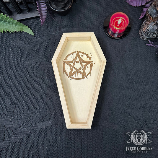 A wooden coffin-shaped tray with a laser-engraved pentagram design, displayed on a black textured fabric background with gothic props including a red candle, a skull, and green leaves.