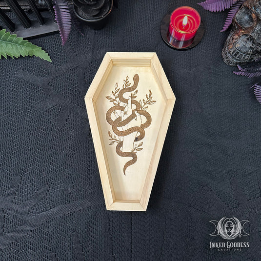 A coffin-shaped wooden tray with a snake and candle design, displayed on a black textured cloth with gothic props including a red candle, skull, and green leaves. Inked Goddess Collection logo is visible in the bottom right corner.