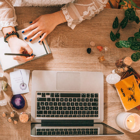 Modern witch at a desk taking notes with a pen and paper next to an open laptop, crystals, plants, and herbs.