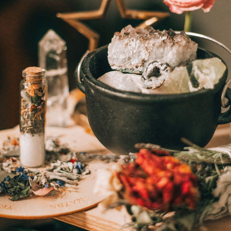 Photo of witchy supplies: small black metal cauldron filled with raw crystals next to a vial of salt and herbs and a bundle of dried flowers.